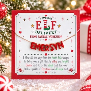 May include: A gold-tone chain necklace featuring red letter beads spelling the name EMERSYN, displayed on a white card with red borders. The card text reads A SPECIAL ELF DELIVERY FROM SANTA'S WORKSHOP and includes a festive holiday poem.
