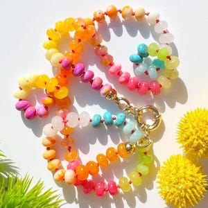 May include: A colorful beaded necklace with a gold clasp. The necklace features a variety of beads in shades of yellow, orange, pink, and turquoise. The beads are strung together with red string, creating a vibrant and eye-catching accessory.