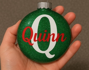 Könnte beinhalten: Ein grüner Glitzer-Weihnachtsschmuck mit dem Namen "Quinn" in roter Schrift über einem großen weißen Buchstaben "Q". Der Schmuck hat eine silberne Metallkappe und eine Schlaufe zum Aufhängen.