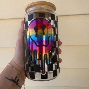 Könnte beinhalten: Ein klares Glas mit einem Bambusdeckel. Das Glas hat ein schwarz-weißes Schachbrettmuster und ein regenbogenfarbenes, schmelzendes Smiley-Gesicht-Design. Das Glas wird von einer Person gehalten.