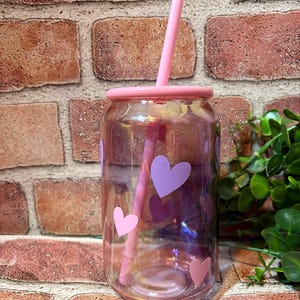 May include: A clear glass tumbler with a pink lid and straw, decorated with pink and purple heart stickers. The iridescent glass reflects light, creating a colorful effect. The tumbler is designed to resemble a soda can.