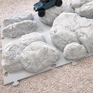 May include: A teal-colored toy SUV is positioned on a gray rock-like playset. The playset is made of interlocking pieces, with a textured surface resembling rocks. The background is a neutral-toned carpet.
