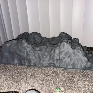 May include: A gray, 3D-printed mountain range model with a textured surface. The model has a base and is designed to resemble a natural rock formation. The color is a solid gray.