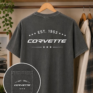 May include: A charcoal grey short-sleeve t-shirt featuring a white graphic on the back that reads EST. 1953 CORVETTE with stars. A circular inset shows a matching pocket on the front with the same white text and star design printed on the fabric.