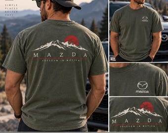 Può includere: Una t-shirt verde oliva con un piccolo logo Mazda sul petto. Il retro mostra una grafica di cime montuose bianche, un sole rosso e il testo Mazda Freedom in Motion. La t-shirt è indossata con un berretto da baseball scuro.