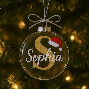 May include: Clear glass Christmas ornament with the name "Sophia" in white script and a gold glitter "S". A small red Santa hat is on the right side of the ornament. The ornament hangs from a silver string and bow.