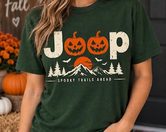 May include: A dark green short-sleeve t-shirt featuring a graphic print with the text Jeep, where the two letter e's are replaced by orange jack-o'-lanterns. Below, a mountain landscape with a sun, pine trees, bats, and the text Spooky Trails Ahead.