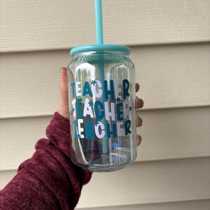 May include: Clear glass tumbler with a teal lid and straw. The tumbler has the word "TEACHER" in teal and white lettering. The tumbler is held in a hand with a maroon sleeve.