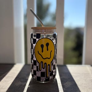 May include: A clear glass tumbler with a black and white checkered pattern and a yellow dripping smiley face design. The tumbler has a bamboo lid and a stainless steel straw.