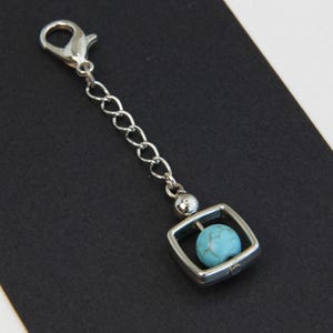 May include: A silver-toned charm with a lobster clasp and chain. The charm features a square frame with a small, round, turquoise-colored bead inside. The background is black and white.