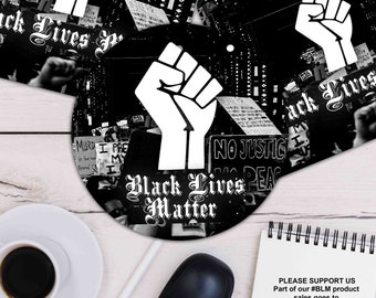 May include: Black and white round mouse pad with a raised fist in the center and the words "Black Lives Matter" in a stylized font. The background is a blurry image of a protest with signs that read "No Justice No Peace" and "Black Lives Matter".