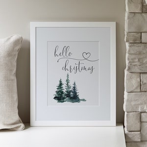 May include: A white framed print with the text "hello christmas" and a watercolor illustration of three green pine trees. The print is hanging on a white wall above a white shelf with a wooden top. There is a deer figurine on the shelf, a glass candle holder with a lit candle, and a small green plant.