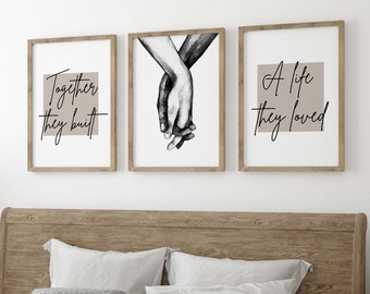 May include: Three framed prints with black calligraphy on a beige background. The prints feature the text "Together they built", "A life they loved", and a black and white illustration of two hands holding each other. The prints are hanging on a white wall above a wooden bed with white bedding and a wooden bench.
