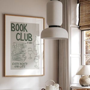 May include: A framed poster with the text "Book Club ( & Wine)" and a drawing of a stack of books, a bottle of wine, and two wine glasses. The poster is hanging on a white wall above a white table with a marble top. Two wicker chairs are positioned on either side of the table.