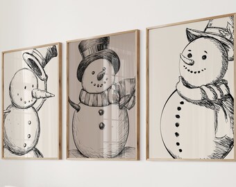 May include: Three framed prints showcasing snowman sketches. Each artwork depicts a snowman with a hat, scarf, and carrot nose. The prints are displayed above a white sofa with cushions and a wooden coffee table, creating a cozy, festive atmosphere.