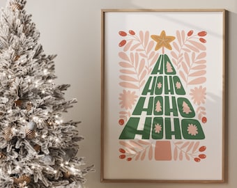May include: A white flocked Christmas tree with gold and white baubles and fairy lights, next to a framed print of a green Christmas tree with the text "HO HO HO" in a pink and green colour scheme. The print is on a white background with pink and red floral accents.