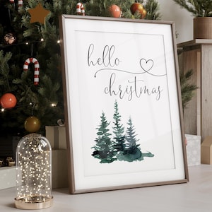 May include: A framed print with the text "hello christmas" in a cursive font with a small heart above the word "christmas". The print features a watercolor illustration of three green pine trees.