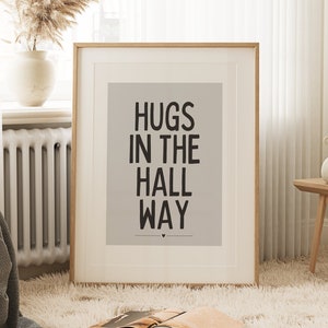May include: A framed print with the words "HUGS IN THE HALL WAY" in black text on a light gray background. The print is in a room with a fluffy white rug, a wooden bench, and a vase with dried flowers.