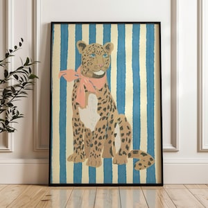 May include: Framed art print of a leopard with a pink scarf, set against blue and gold vertical stripes. The leopard has brown spots and blue eyes. The artwork is displayed on a wooden floor. The frame is black.