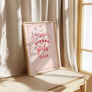 Op de afbeelding: Een ingelijst print met een roze achtergrond en een witte omtrek. De tekst "Strong girls club" is geschreven in rode cursieve letters.