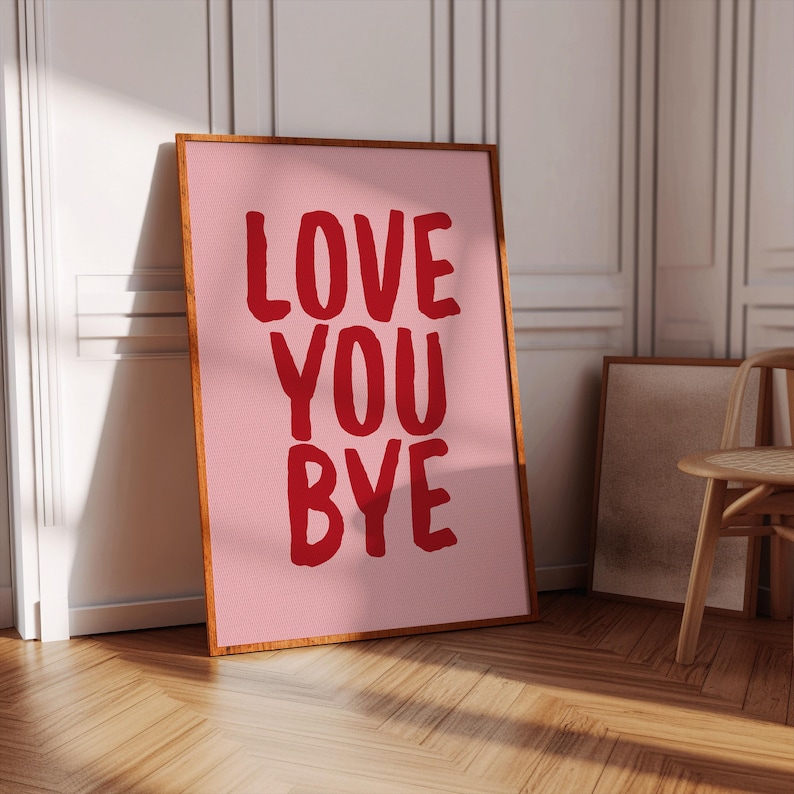 May include: A framed pink art print with the words "LOVE YOU BYE" in bold red letters. The artwork is displayed against a white wall and wooden floor, with a wooden frame. A chair and another framed print are in the background.