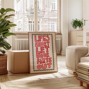 May include: A framed art print with the text "LIFES SHORT LET YOUR FARTS OUT" in red letters. The print has a light pink background and a green scalloped border. The frame is a light brown color.