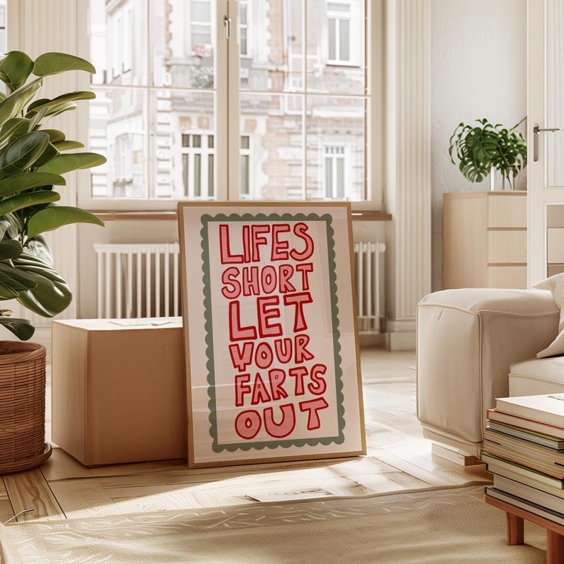 May include: A framed art print with the text "LIFES SHORT LET YOUR FARTS OUT" in red letters. The print has a light pink background and a green scalloped border. The frame is a light brown color.