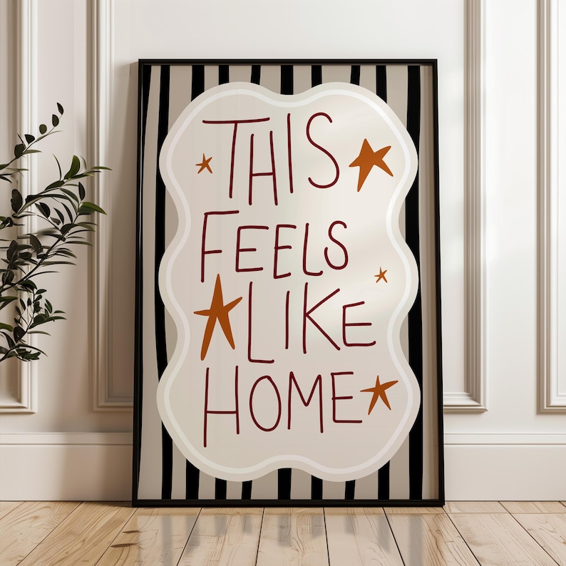 Puede incluir: Impresi&oacute;n art&iacute;stica enmarcada con las palabras "THIS FEELS LIKE HOME" en escritura burdeos. El texto est&aacute; sobre una forma crema con bordes ondulados, acentuada con estrellas naranjas. El fondo presenta rayas verticales blancas y negras.