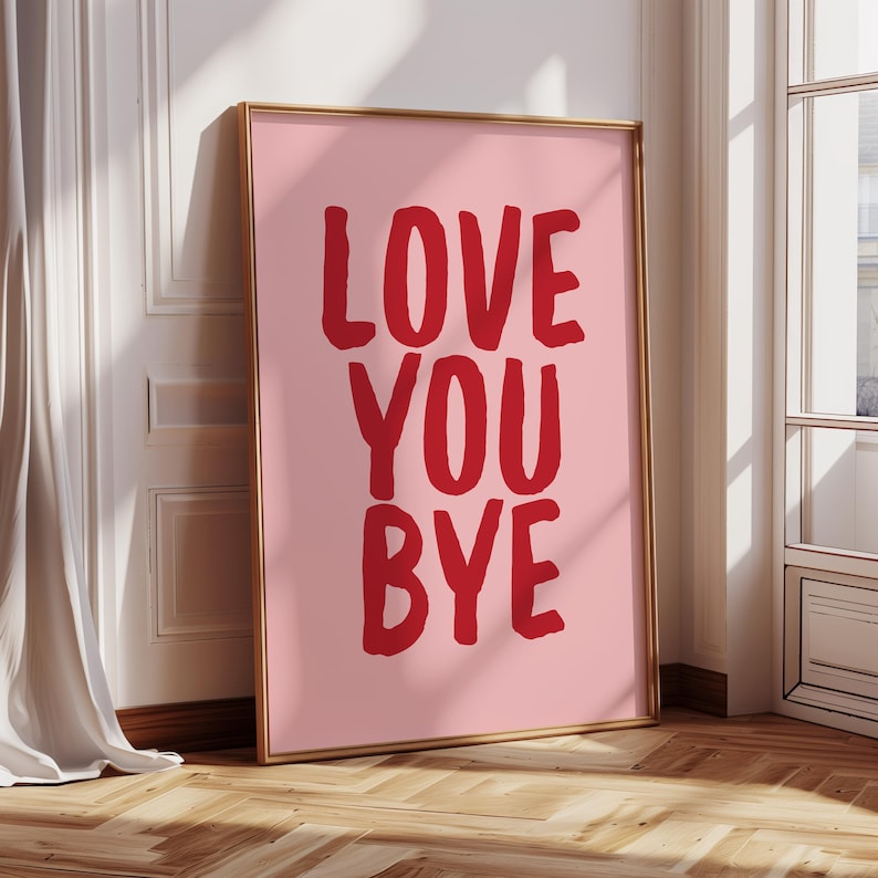 May include: A framed pink art print with the words "LOVE YOU BYE" in bold red letters. The artwork is displayed in a gold frame and leans against a white wall. The room has a wooden floor and a white curtain.