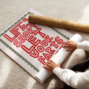 May include: A poster with the text "LIFE'S SHORT LET YOUR FARTS" in red letters. The poster is being unrolled, revealing the message. A rolled-up poster is also visible. The poster has a scalloped green border.