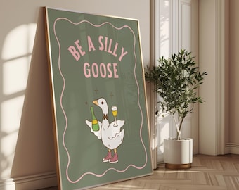 Peut inclure: Une affiche encadrée avec une illustration d'oie de dessin animé tenant une bouteille de champagne et un verre de champagne. L'affiche porte le texte "BE A SILLY GOOSE" en rose sur un fond vert, avec un bord rose. L'oie porte des baskets roses.