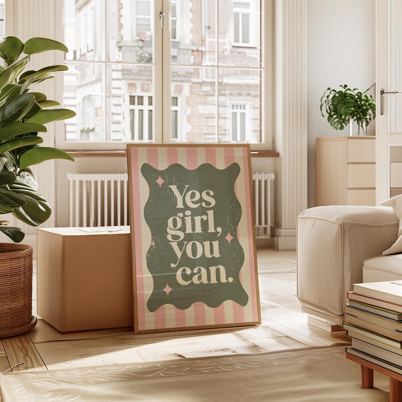 May include: A framed poster with the text "Yes girl, you can." The poster has a green, wavy design with pink stripes and small pink star accents. The poster is displayed in a well-lit room with a window.