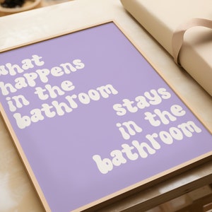 May include: A framed print with a light purple background and white text that reads "What happens in the bathroom stays in the bathroom".