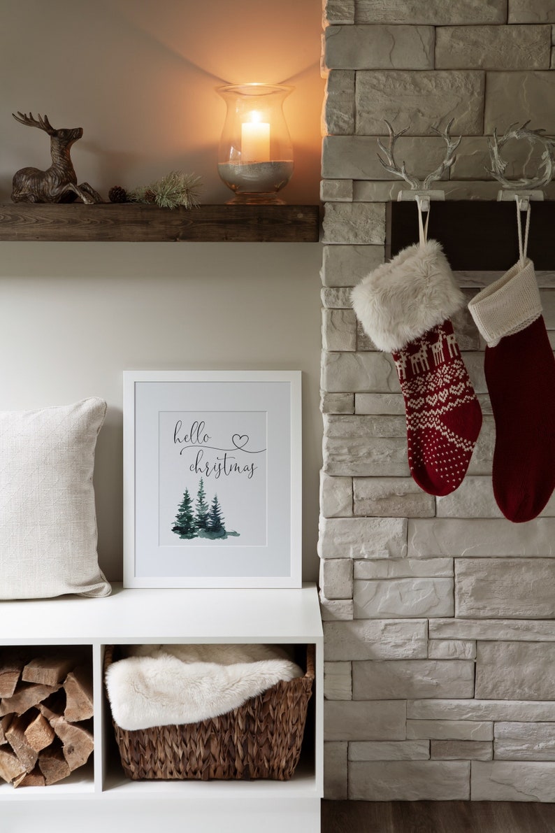 May include: A white framed print with the text "hello christmas" and a watercolor illustration of three green pine trees. The print is hanging on a white wall above a white shelf with a wooden top. There is a deer figurine on the shelf, a glass candle holder with a lit candle, and a small green plant.