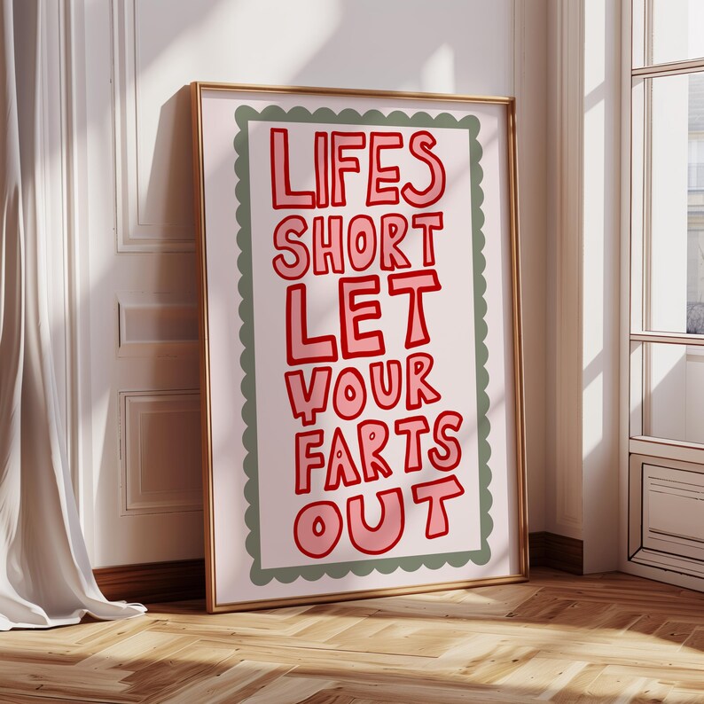 May include: A framed art print with the text "LIFES SHORT LET YOUR FARTS OUT" in red bubble letters. The text is on a light pink background with a green scalloped border, set in a gold frame. The artwork leans against a wall.