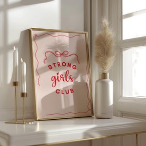 Op de afbeelding: Een gouden ingelijste print met een roze achtergrond en rode tekst die "Strong girls club" luidt met een klein hartje boven het woord "girls".