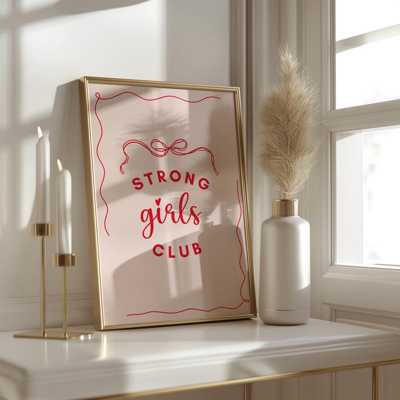 Op de afbeelding: Een gouden ingelijste print met een roze achtergrond en rode tekst die "Strong girls club" luidt met een klein hartje boven het woord "girls".