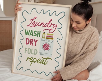 May include: A framed laundry room sign with the words "Laundry, Wash, Dry, Fold, Repeat" in a mix of colors. The sign includes a washing machine graphic and a wavy teal border. The frame is a light wood color.