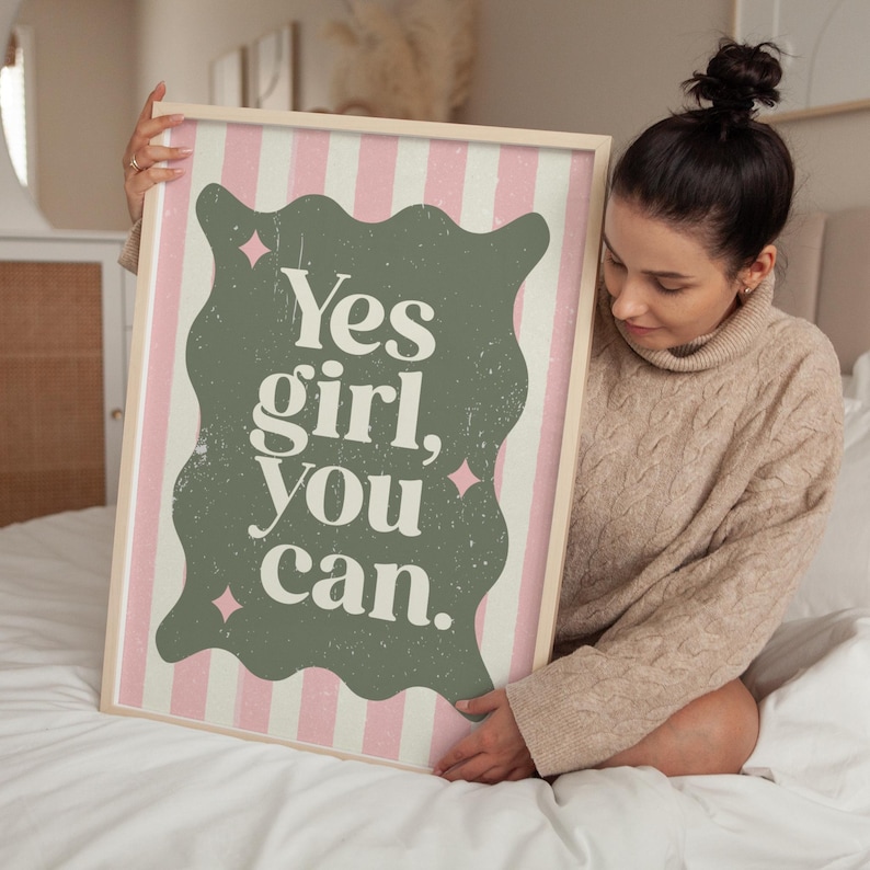 May include: A framed art print with the text "Yes girl, You can." The artwork features a green, wavy-edged shape with the text in white, set against a pink and white striped background. The frame is a light wood color.