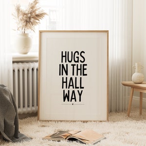 May include: A framed print with the words "HUGS IN THE HALL WAY" in bold black letters. The print is in a light wood frame and is displayed on a fluffy white rug. A vase with dried flowers sits on a windowsill in the background.