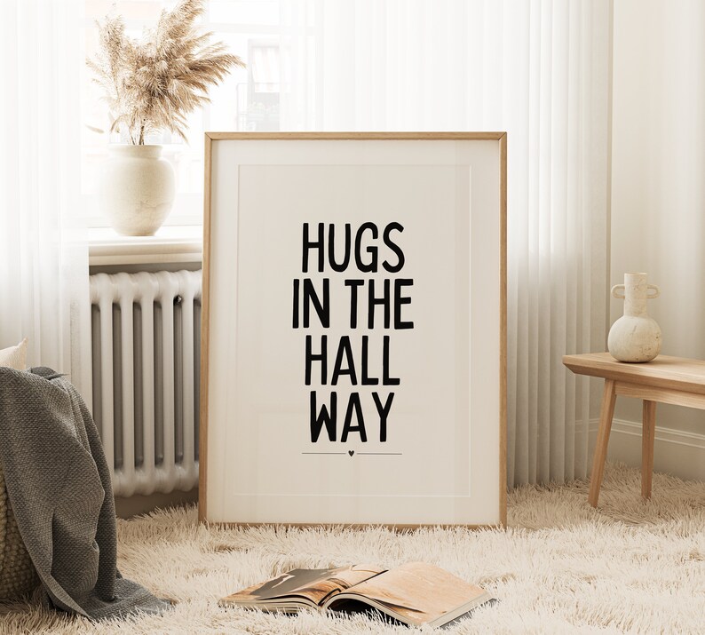 May include: A framed print with the words "HUGS IN THE HALL WAY" in bold black letters. The print is in a light wood frame and is displayed on a fluffy white rug. A vase with dried flowers sits on a windowsill in the background.