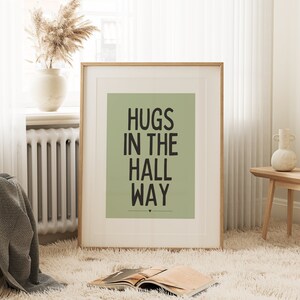 May include: A framed art print with the text "HUGS IN THE HALL WAY" in black on a sage green background. The print is in a light wood frame and is displayed in a cozy room setting with a fluffy rug and decorative items.