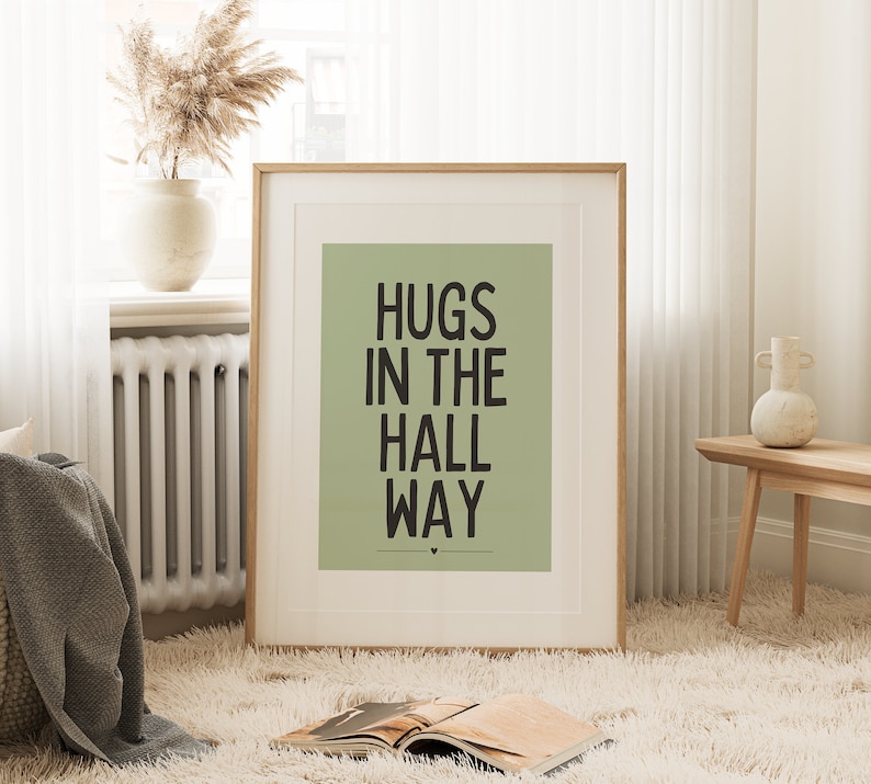 May include: A framed art print with the text "HUGS IN THE HALL WAY" in black on a sage green background. The print is in a light wood frame and is displayed in a cozy room setting with a fluffy rug and decorative items.
