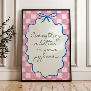 Może przedstawiać: Oprawiony wydruk z napisem "Everything is better in your pyjamas". Grafika przedstawia r&oacute;żowo-białe kratkowane tło, biały owal z niebieską obw&oacute;dką i niebieską kokardą. Rama jest czarna.