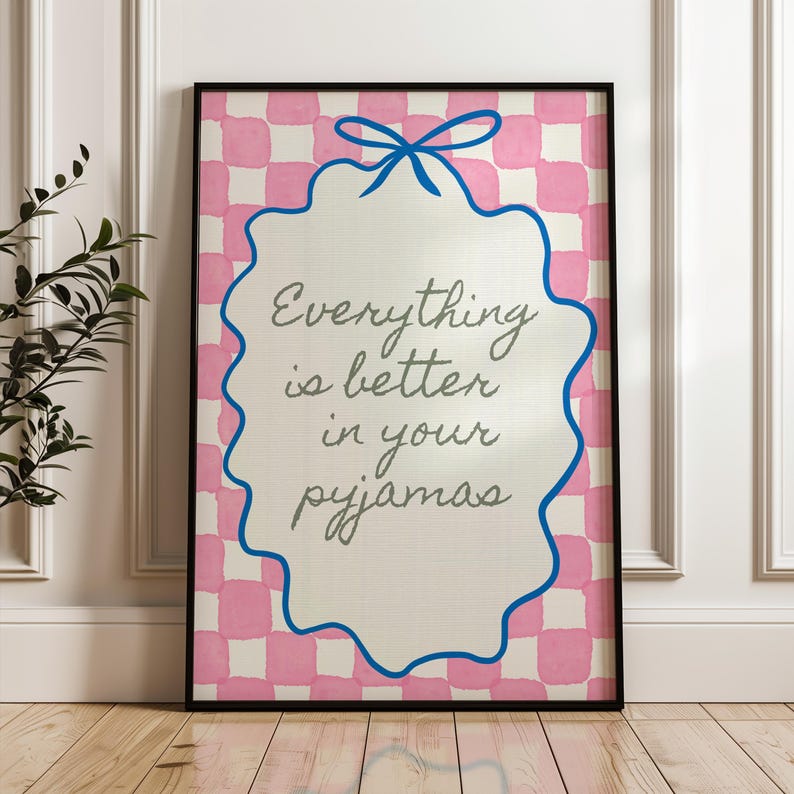 Może przedstawiać: Oprawiony wydruk z napisem "Everything is better in your pyjamas". Grafika przedstawia r&oacute;żowo-białe kratkowane tło, biały owal z niebieską obw&oacute;dką i niebieską kokardą. Rama jest czarna.