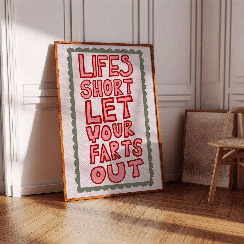 May include: A framed art print with the text "LIFES SHORT LET YOUR FARTS OUT" in red, outlined in white, on a white background. The print has a green scalloped border and a wooden frame. The artwork leans against a white wall.