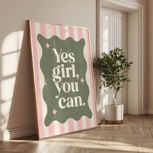 May include: A framed art print with a gold border, leaning against a wall. The print features a pink and white striped background with a green, wavy-edged shape in the center. The words "Yes girl, you can" are printed in white.