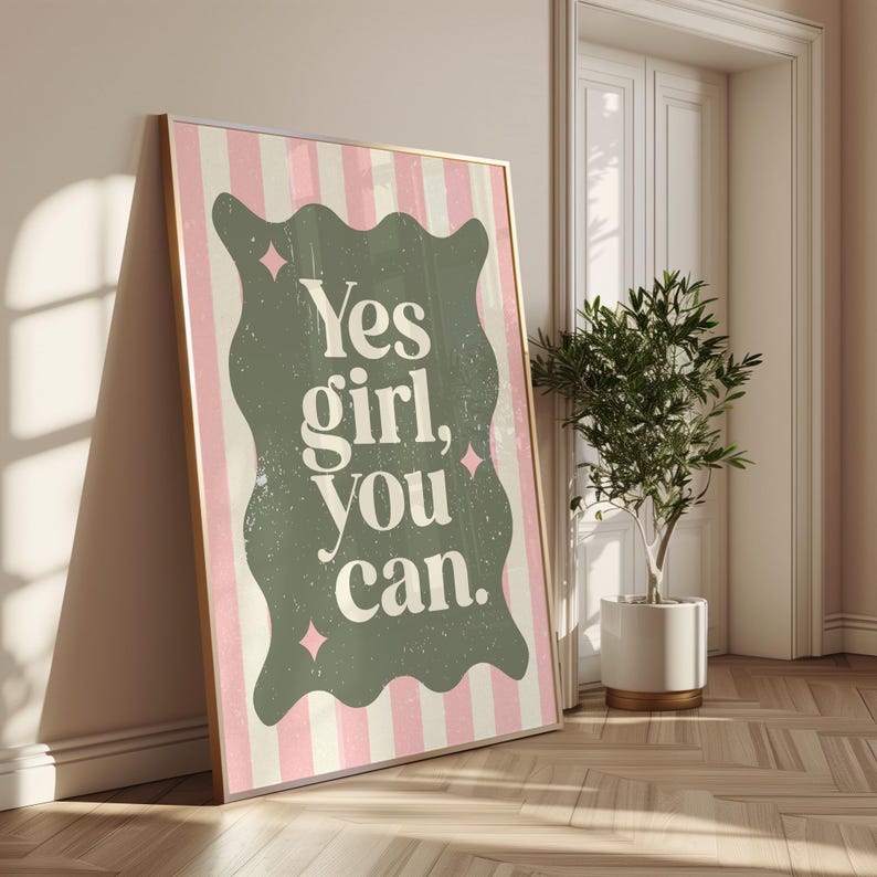 May include: A framed art print with a gold border, leaning against a wall. The print features a pink and white striped background with a green, wavy-edged shape in the center. The words "Yes girl, you can" are printed in white.