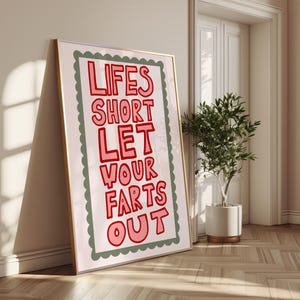 May include: A framed art print with the text "LIFES SHORT LET YOUR FARTS OUT" in red letters on a white background. The print has a scalloped green border and a gold frame, leaning against a wall.