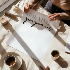 May include: A white card with the text "Morning gorgeous" written in black cursive script. The card is lying on a white surface with a cup of coffee and a shadow.